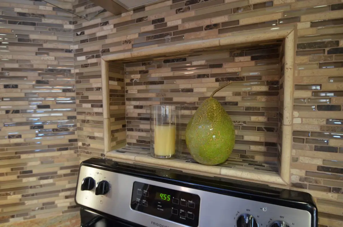 “Kitchen backsplash niche with decorative tile surround, glass candle holder, and ceramic pear above stove.”