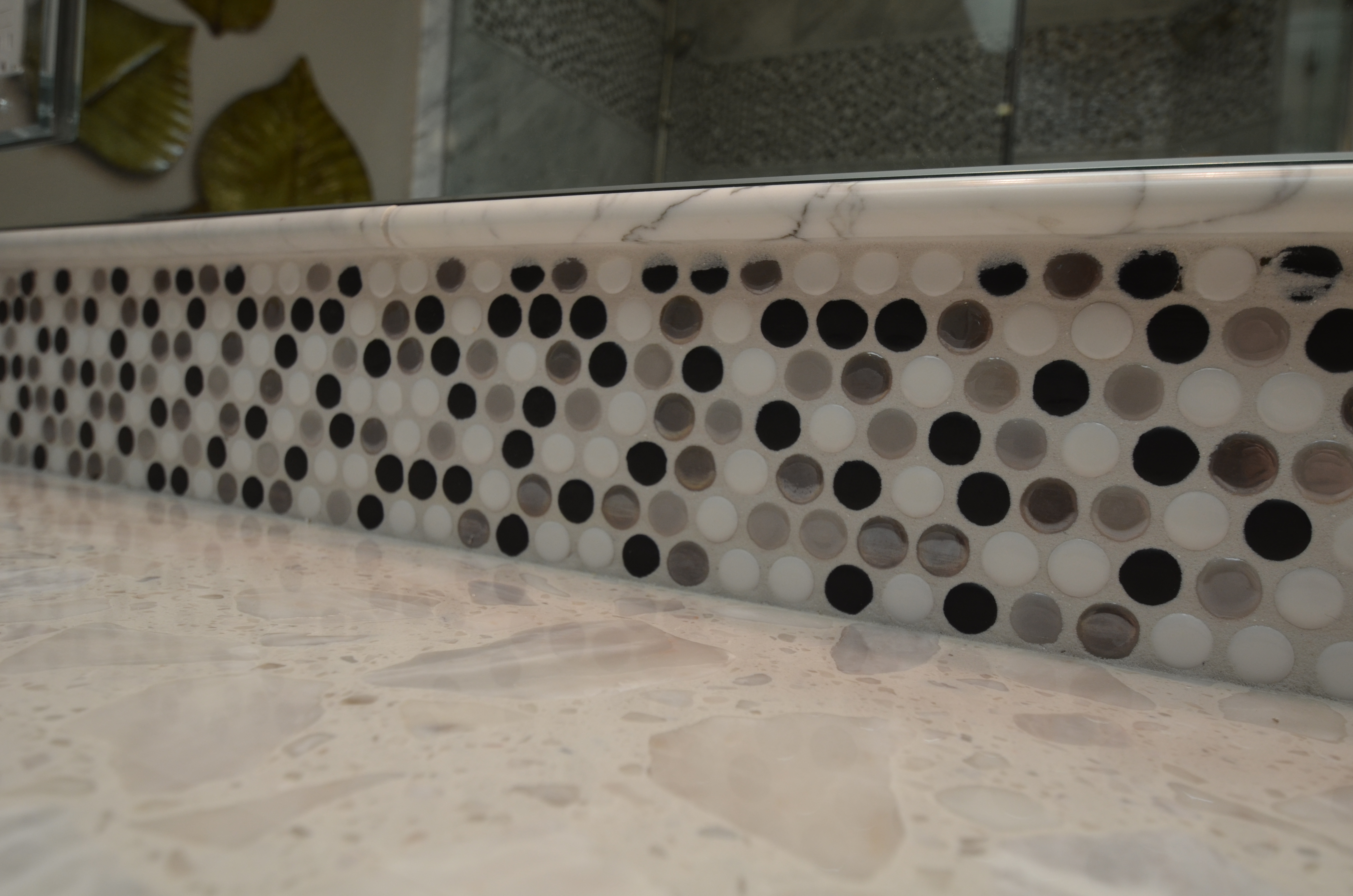 Close-up of a bathroom backsplash featuring circular mosaic tiles in shades of white, gray, and black above a polished stone countertop.