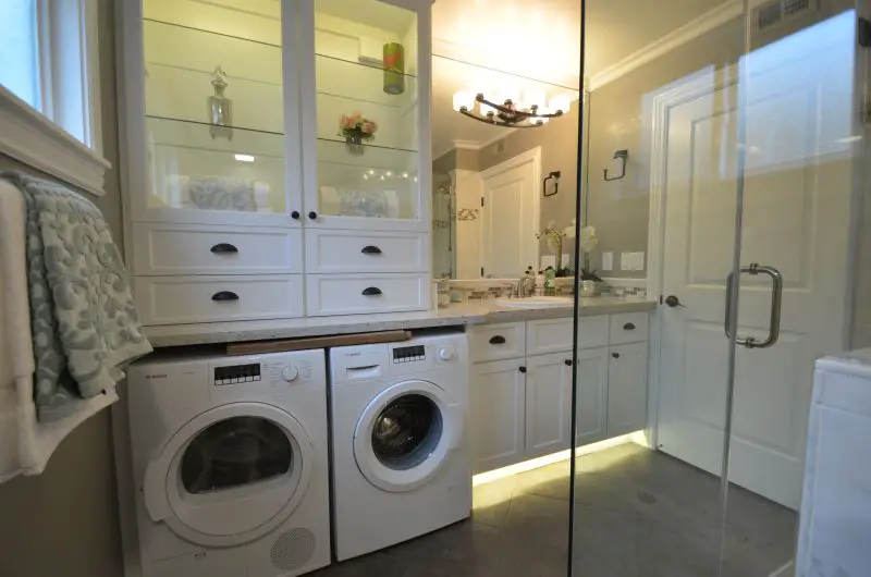 Modern bathroom with stacked storage cabinets, built-in vanity, and side-by-side front-loading washer and dryer beneath the counter. Glass shower enclosure, elegant lighting, and soft gray tones create a clean and functional design.