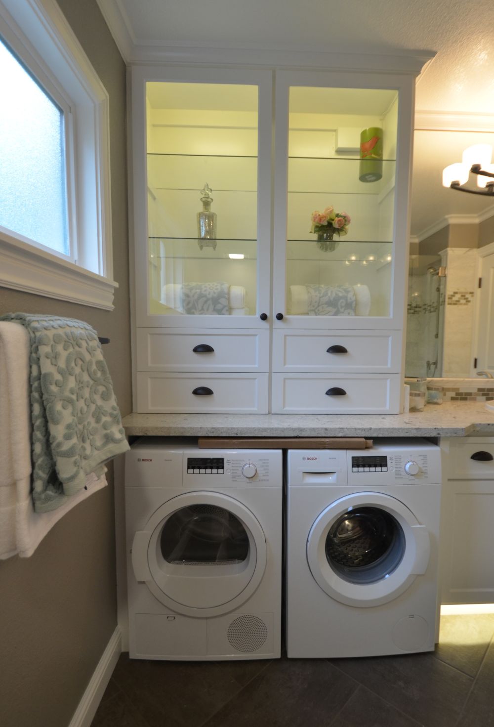 Side-by-side front-loading washer and dryer installed beneath a stone countertop with custom glass-front cabinets above, forming a built-in laundry nook within a bathroom setting in a Rossmoor home that maximizes storage while maintaining a clean, cohesive design.