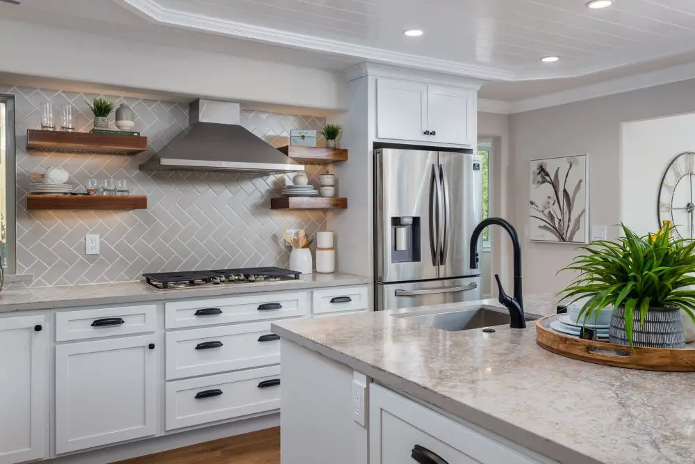 White shaker kitchen with quartz countertops, floating wood shelves, stainless steel range hood, and herringbone tile backsplash.