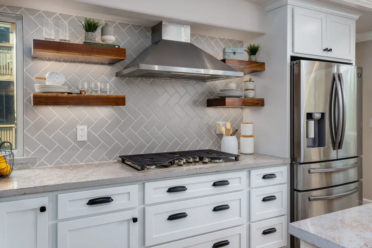 Contemporary kitchen featuring white cabinetry, light gray herringbone tile backsplash, floating wood shelves with accent lighting, stainless steel range hood, and built-in refrigerator.