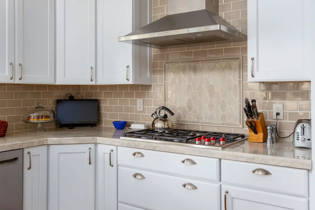 Traditional kitchen with white cabinets, beige subway tile backsplash, and a decorative hexagon tile inset above the stovetop. Stainless steel range hood, marble-look countertops, and gas cooktop complete the space.