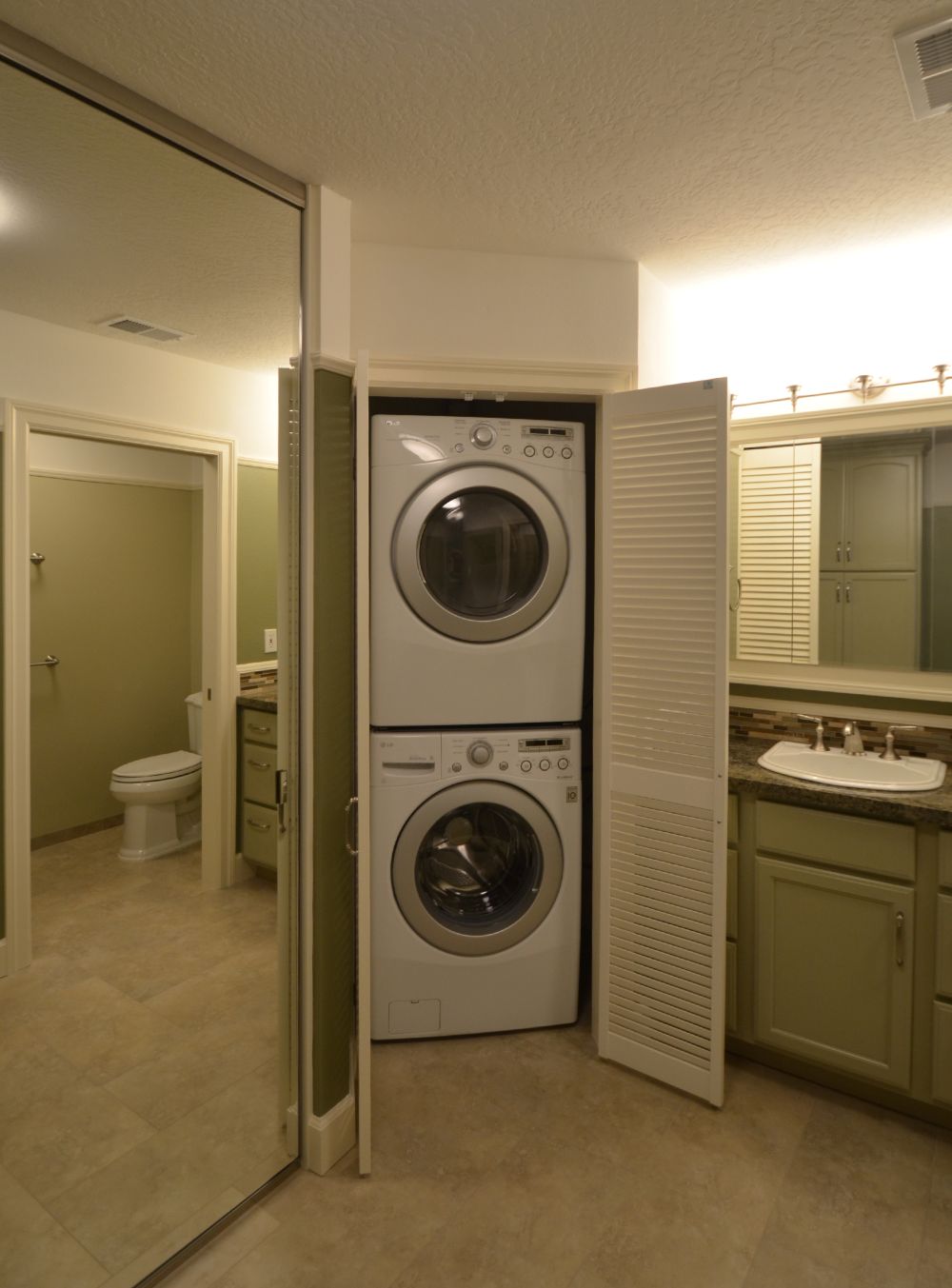 Stacked front-loading washer and dryer installed inside a narrow closet next to a bathroom vanity, featuring louvered doors and a space-saving Rossmoor laundry reconfiguration designed for efficient use of limited square footage.