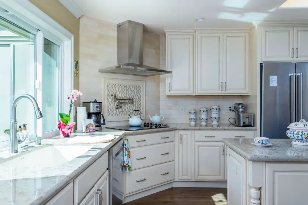 White traditional kitchen cabinets with stone countertops, stainless steel range hood, large window, and center island in a remodeled kitchen.