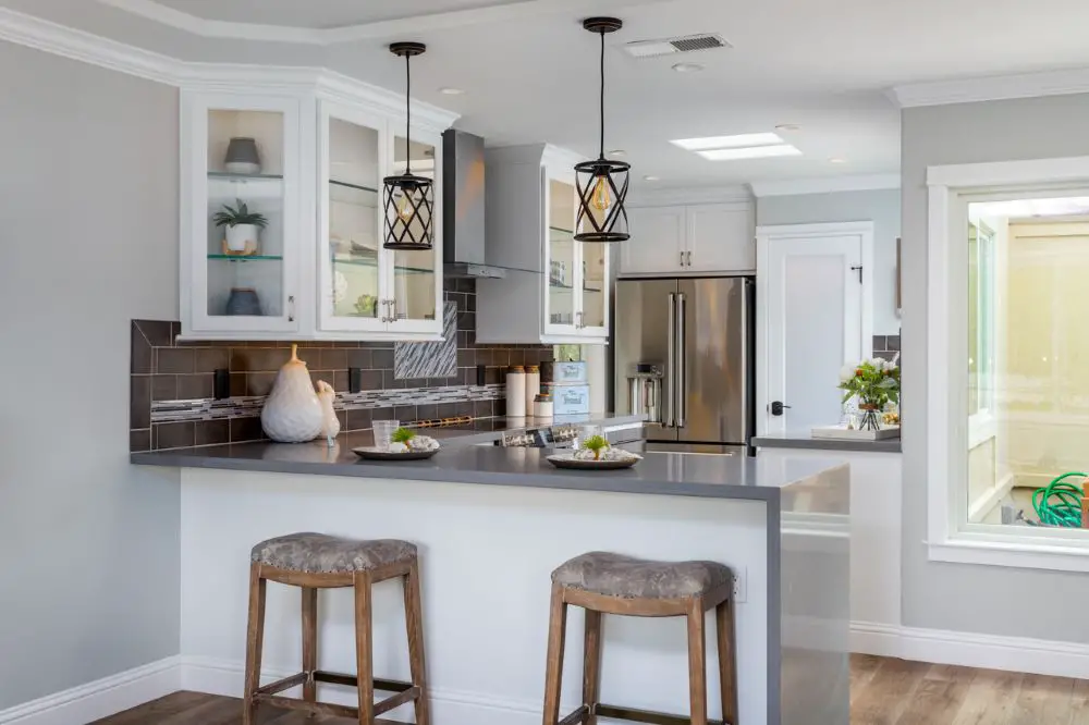 “Light gray quartz countertops in a white transitional kitchen with brown tile backsplash and black pendant lighting.”