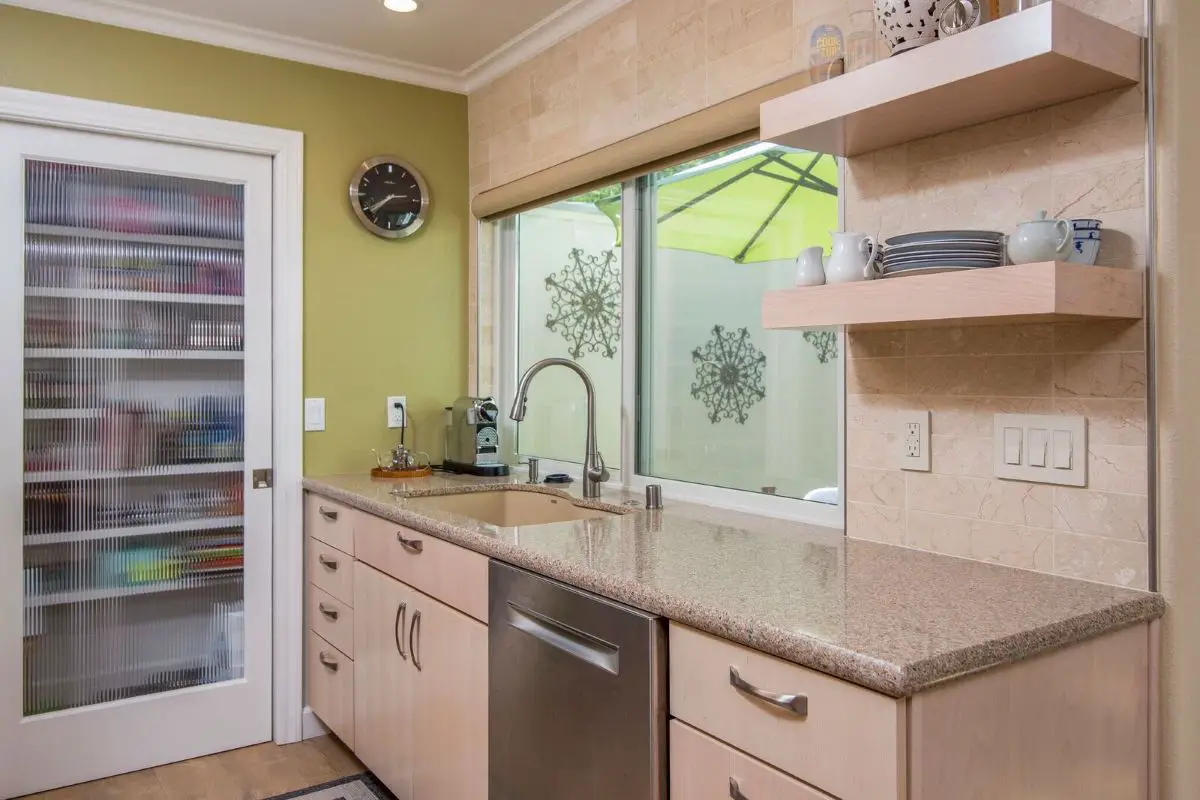 Contemporary kitchen with light wood cabinets, granite countertops, and beige stone tile backsplash. Stainless steel dishwasher and modern faucet beneath a large window, with open wood shelves holding dishes and mugs. Green accent wall with frosted glass pantry door on the left.