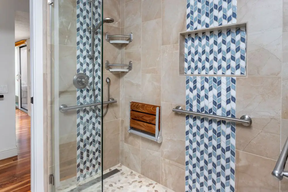 Accessible shower with fold-down teak bench, dual grab bars, and blue geometric mosaic accent tiles — stylish aging-in-place remodel.