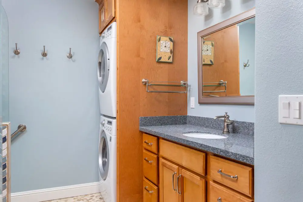 Compact bathroom with a stacked washer and dryer built into a wooden cabinet beside a single-sink vanity with granite countertop and light blue walls.