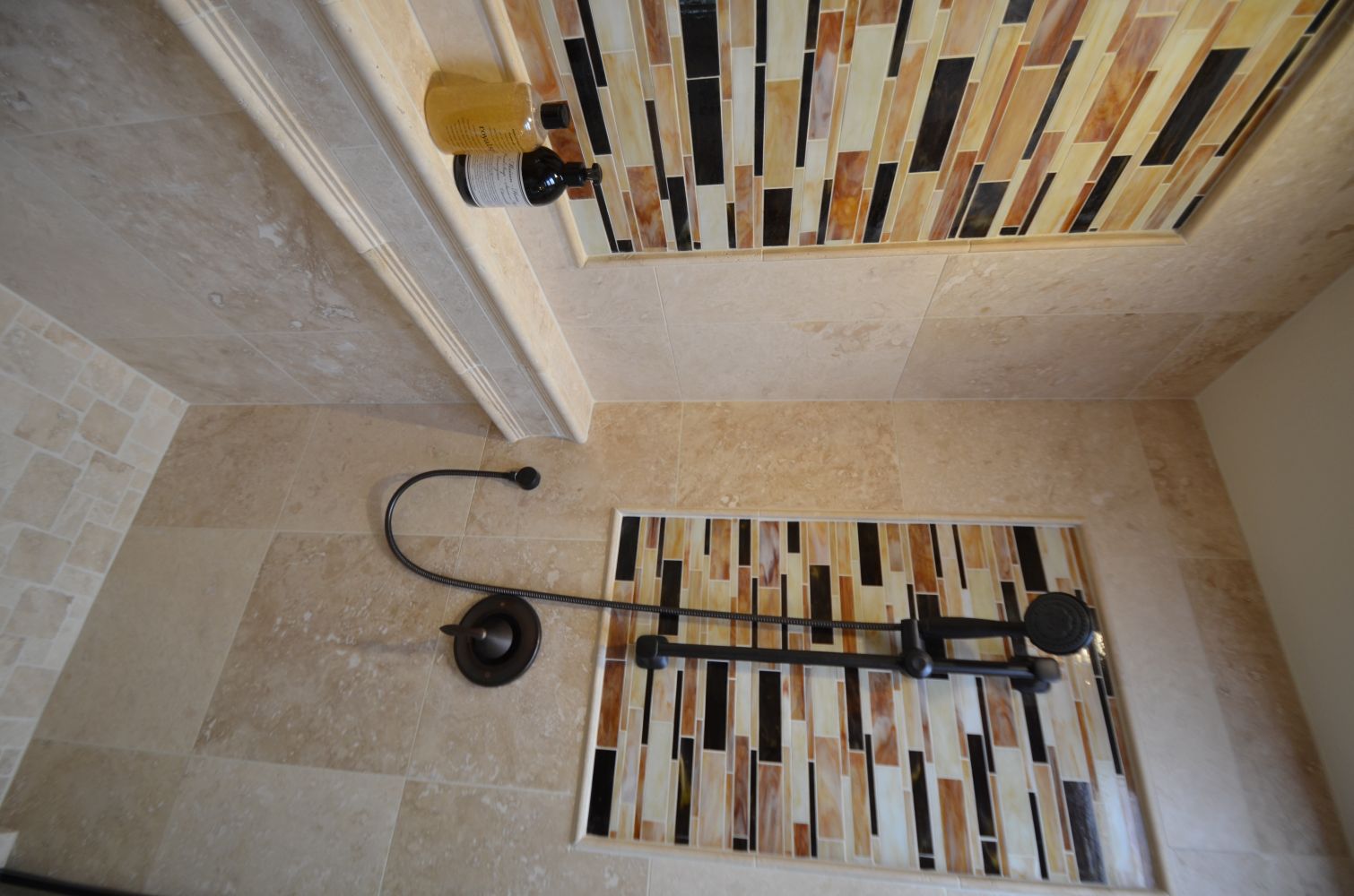Warm travertine tile shower with rectangular amber, cream, and black glass mosaic insets. Oil-rubbed bronze shower fixture and handheld sprayer mounted on a slide bar.