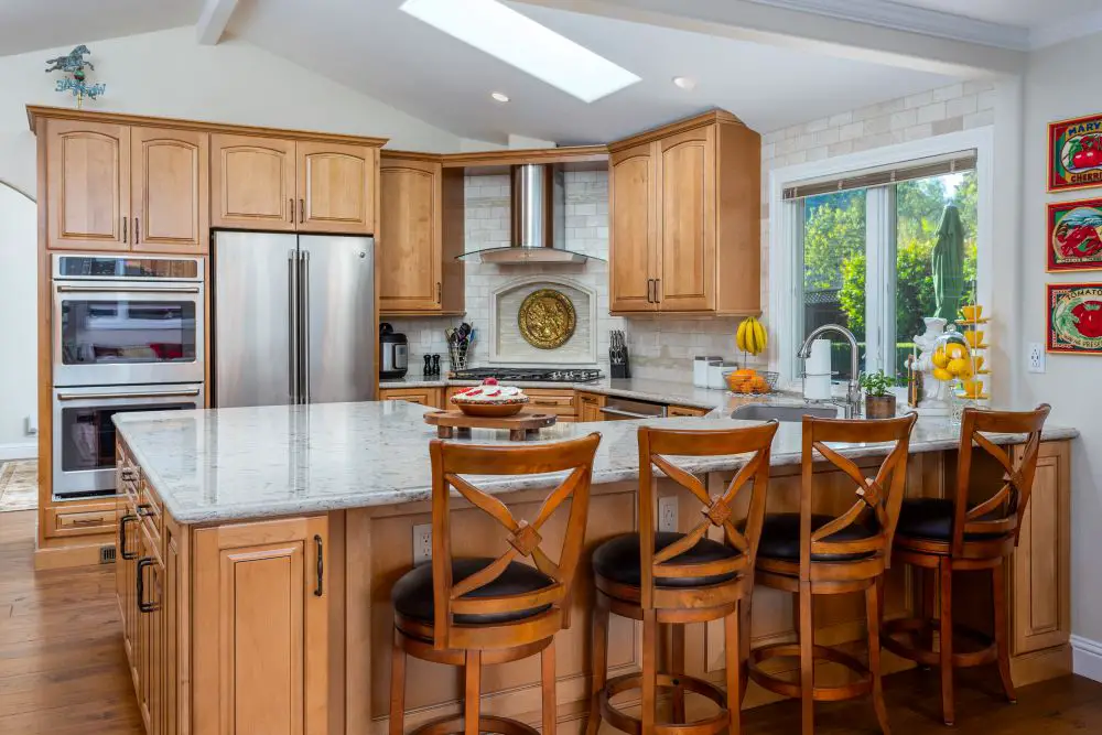 Open-concept kitchen with warm maple cabinetry, quartz countertops, and large island with bar seating. Skylight and sliding glass door fill the space with natural light, highlighting the custom backsplash and stainless steel range hood.