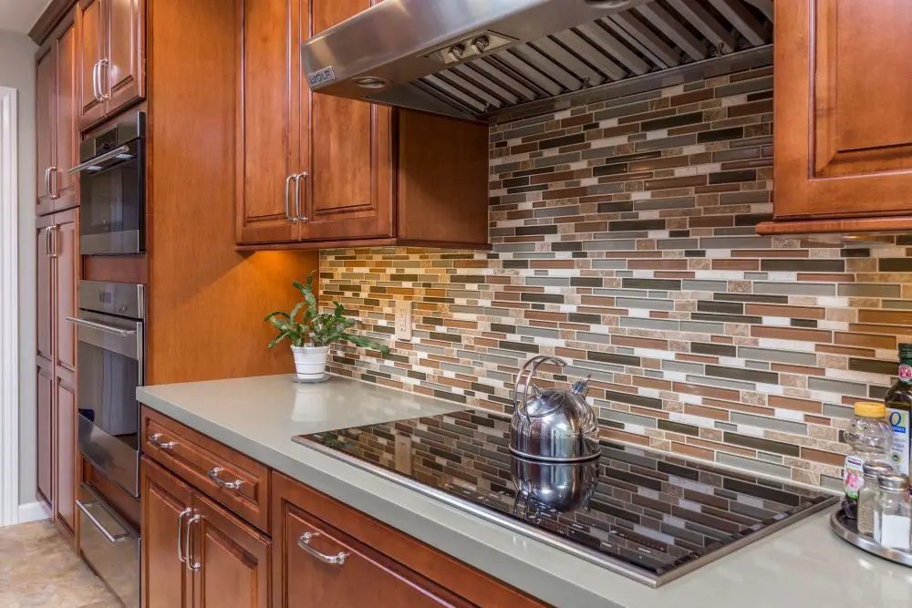 “Soft gray quartz countertop in a warm cherry wood kitchen with a multi-tone mosaic tile backsplash and electric cooktop.”