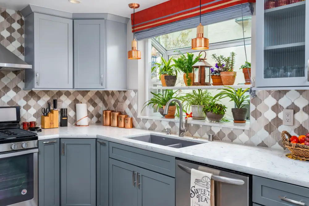 Gray shaker kitchen cabinets with patterned tile backsplash, quartz countertops, farmhouse sink, and large window filled with houseplants.