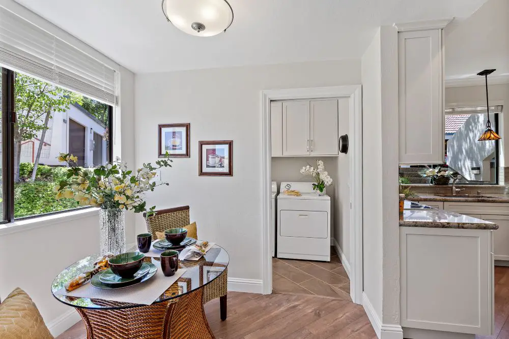 Sunlit dining area with a round glass table set for breakfast, adjacent to a compact laundry space with a white dryer and overhead cabinets.