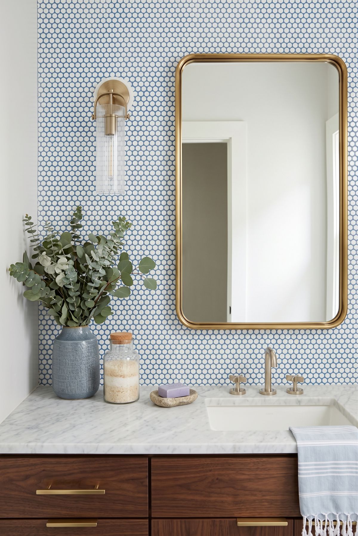 A close-up view of a modern-traditional bathroom vanity featuring vibrant blue penny round tile wallpaper, a polished gold rectangular mirror, and a ribbed glass wall sconce.