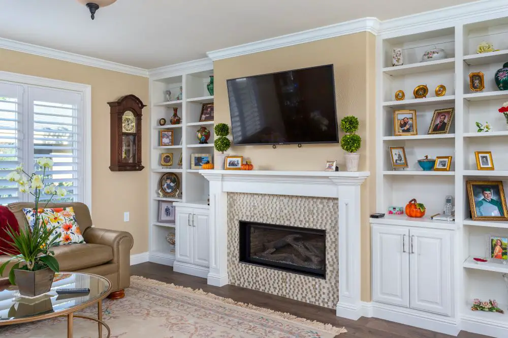 White built-in bookshelves and cabinets flanking a mosaic tile fireplace with wood mantel and wall-mounted TV in a beige living room