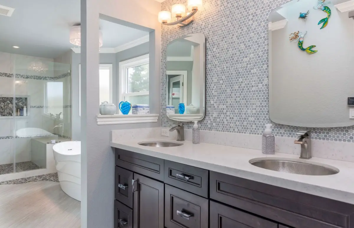 Interior view of a master bathroom remodel showcasing a grey patterned mosaic tile backsplash, dark espresso double vanities with white quartz counters, and a freestanding tub in an adjacent wet room.