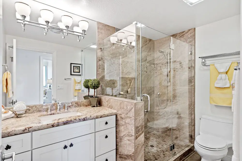 Bathroom with beige stone tile walk-in shower, glass enclosure, pebble floor, and double vanity with granite countertop