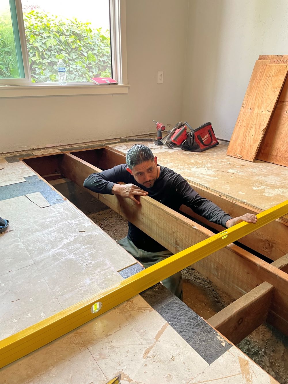 Worker repairing subfloor framing using level tool during home renovation.