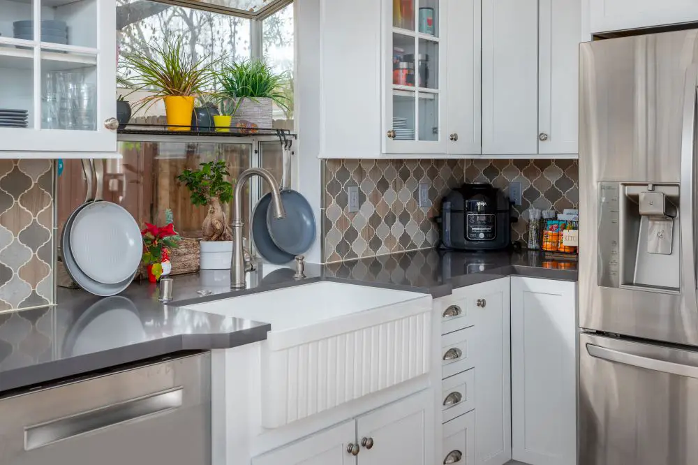Gray quartz countertops surrounding a white farmhouse sink, with white cabinets and a patterned tile backsplash.