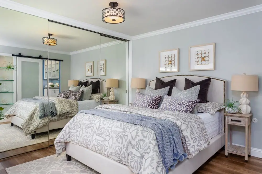 Soft gray bedroom with mirrored closet doors, upholstered bed, layered bedding, and modern ceiling light fixture.