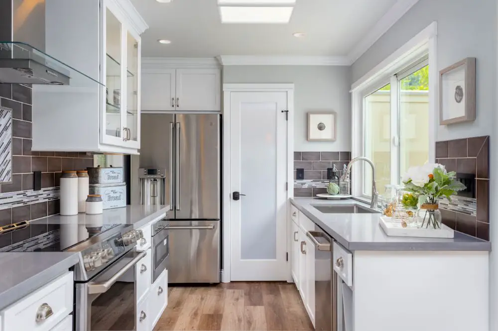 Bright galley kitchen with white shaker cabinets, gray subway tile backsplash, stainless steel appliances, and a skylight providing natural light.