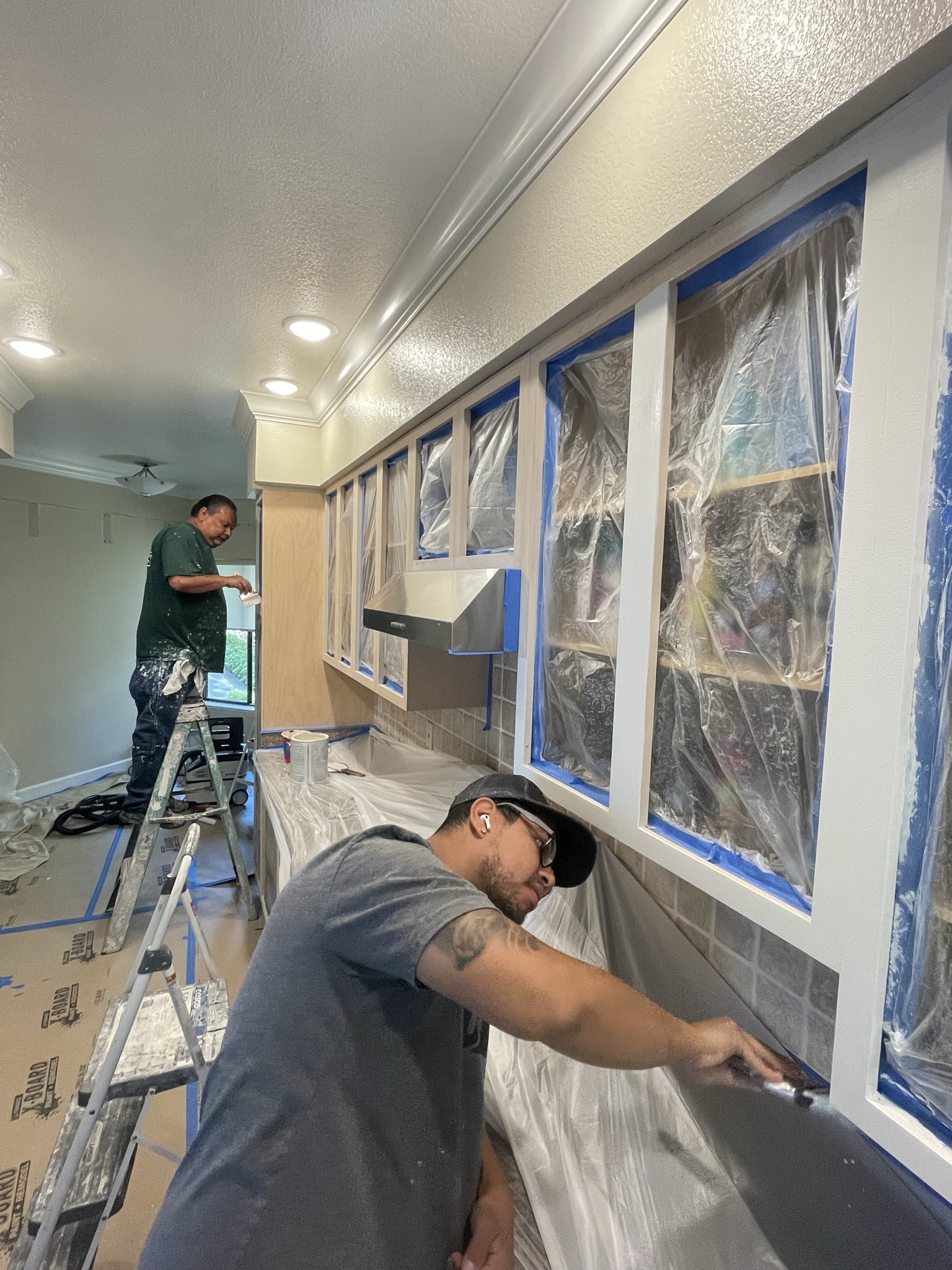 Workers painting kitchen cabinets with plastic covering and masking for refinishing.