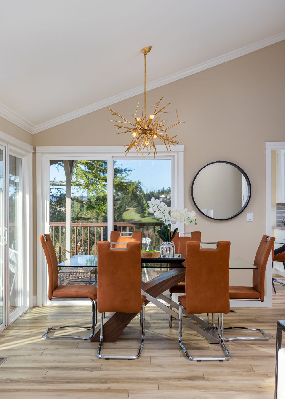 Modern dining room with glass table, caramel leather chairs, gold chandelier, large windows, and scenic outdoor views.