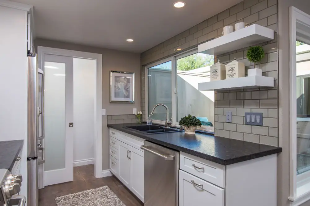 Floating open shelves above a kitchen sink with gray subway tile backsplash, white cabinets, and stainless steel appliances.