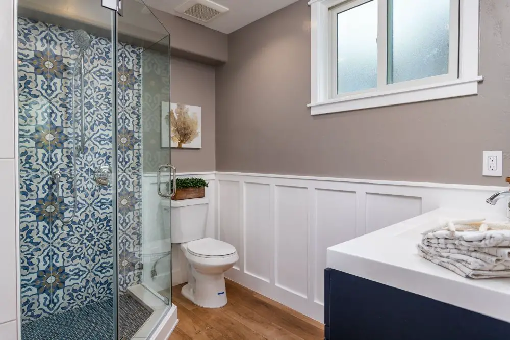 Bathroom with blue patterned tile shower, white wainscoting, glass enclosure, and wood-look flooring
