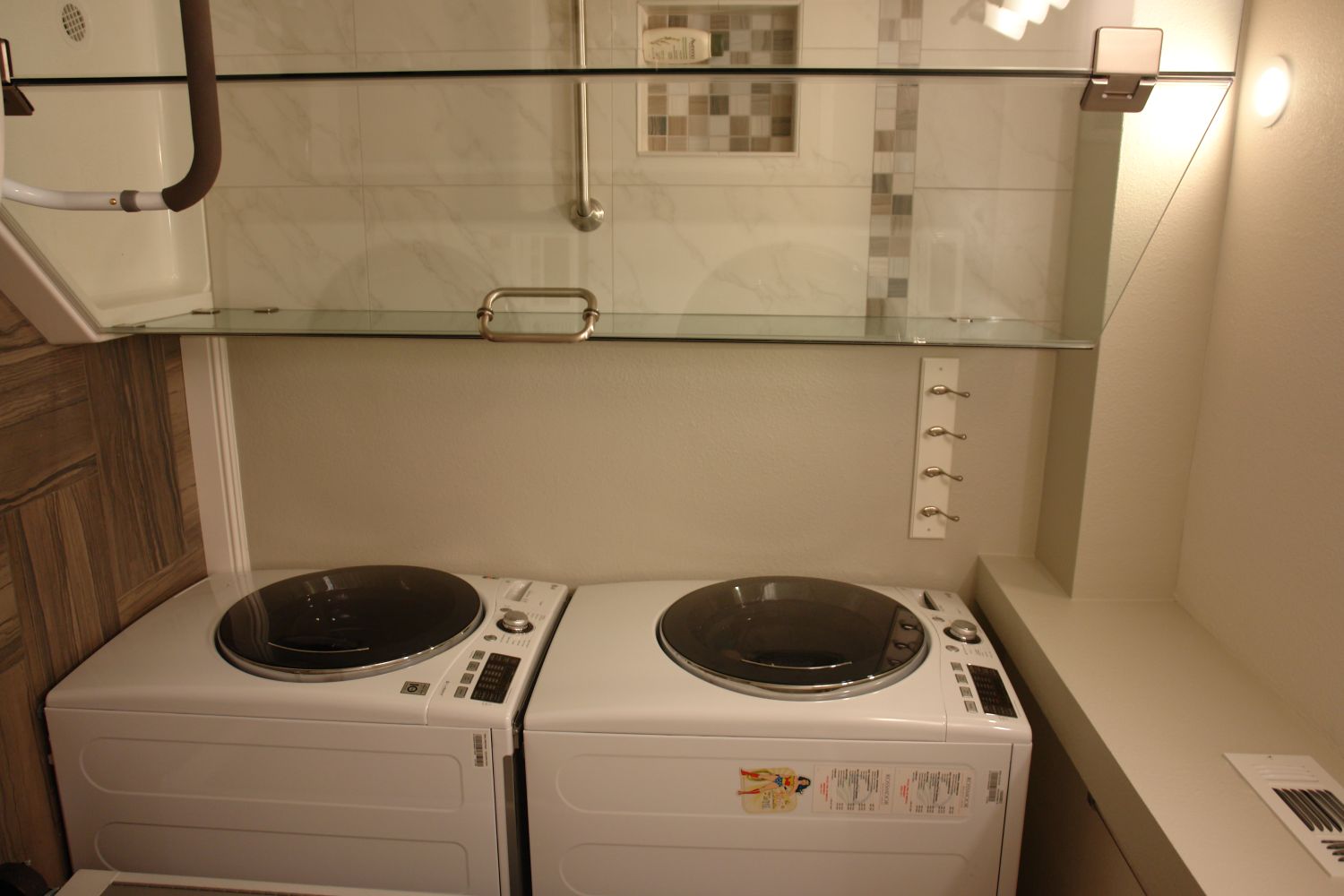 Stacked front-load washer and dryer installed in a Rossmoor bathroom next to a glass-enclosed walk-in shower, with a coat hook rail and open shelf above — showing how in-unit laundry fits alongside existing bathroom features in a Walnut Creek condo remodel.