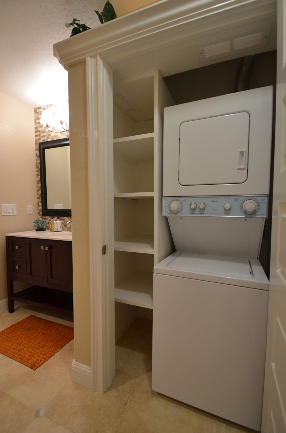 Stacked washer and dryer built into a recessed closet niche beside a bathroom vanity in a Rossmoor condo, with open white shelving on one side for storage — demonstrating a compact, functional laundry retrofit in a Walnut Creek condominium remodel.
