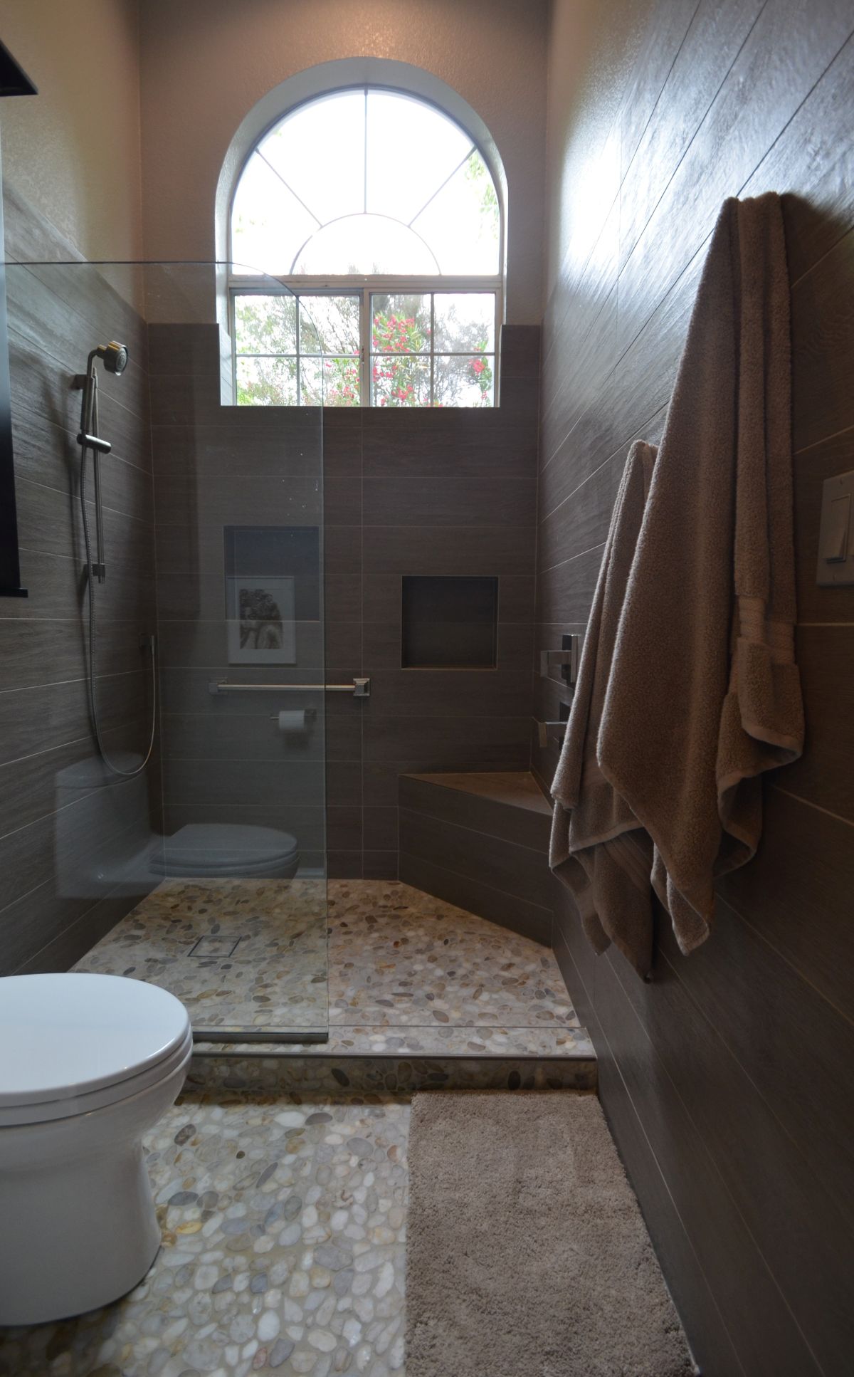 “Low-threshold shower with pebble tile floor, frameless glass enclosure, built-in bench, and large arched window in bathroom remodel.”