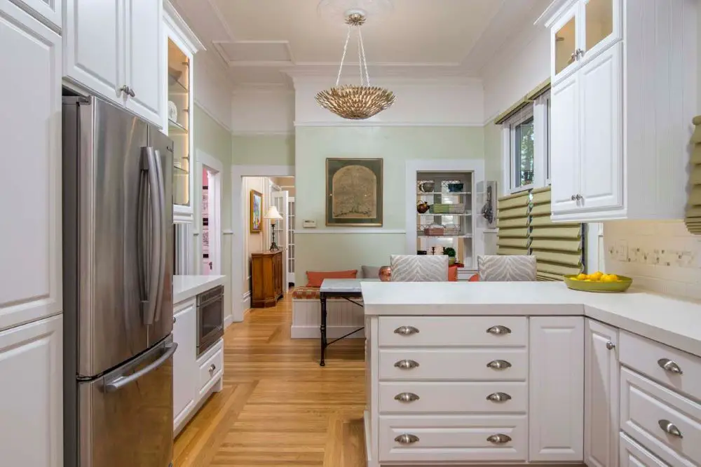 White shaker galley kitchen remodel with quartz countertops, stainless steel appliances, custom cabinetry, hardwood floors, tall ceilings, and a peninsula opening to the living area.